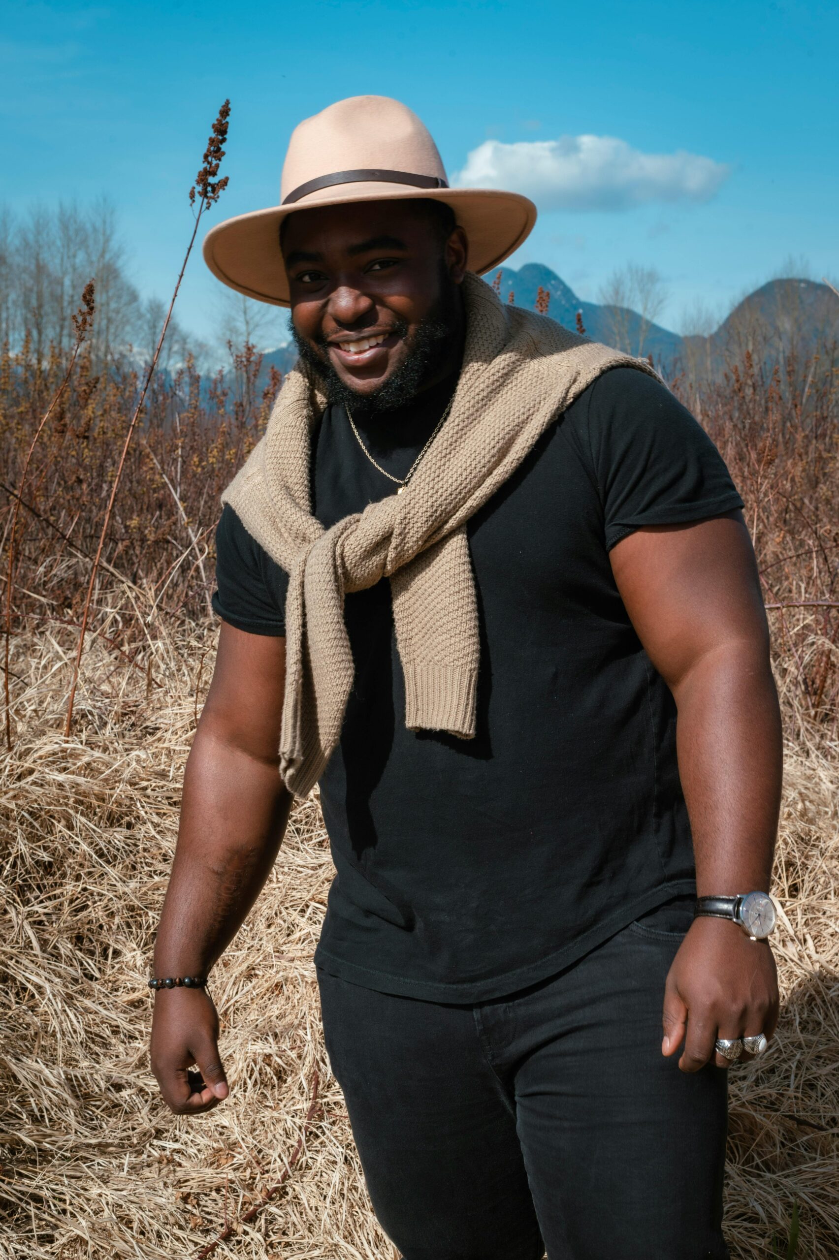 A smiling man in a hat and sweater poses outdoors in a grassy field beneath a blue sky.